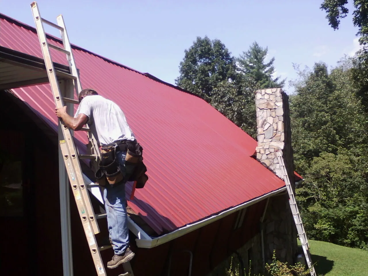 Roofer on a ladder beside a red metal roof and stone chimney — Soffit & Fascia Repair by Roofing Brothers in East Pittsburgh